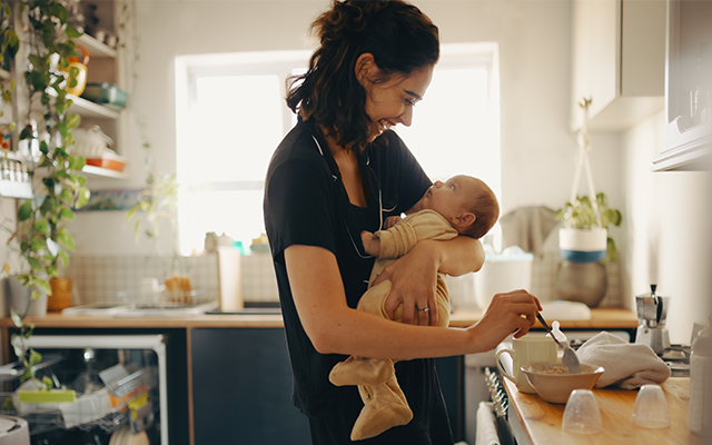 Person holding and feeding an infant in a home kitchen setting.